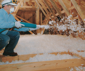 Masked worker installing blown-in fiberglass in an attic.