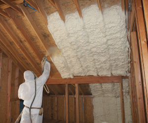 Worker in a white protective suit installing spray foam insulation on an attic ceiling and wall.