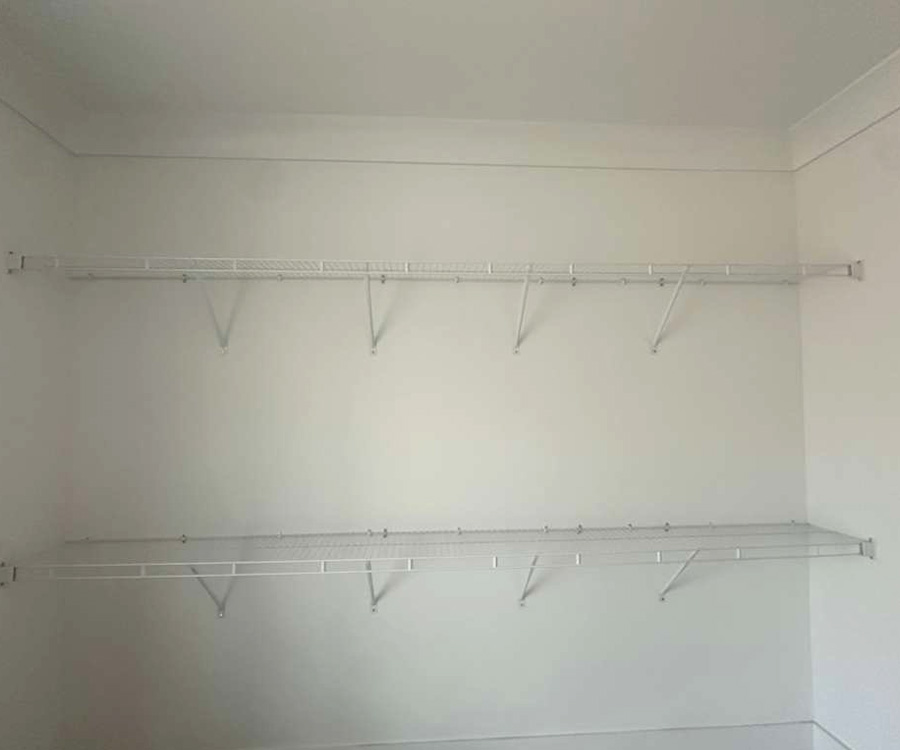 Two levels of empty white wire shelving mounted on a plain white wall with metal brackets